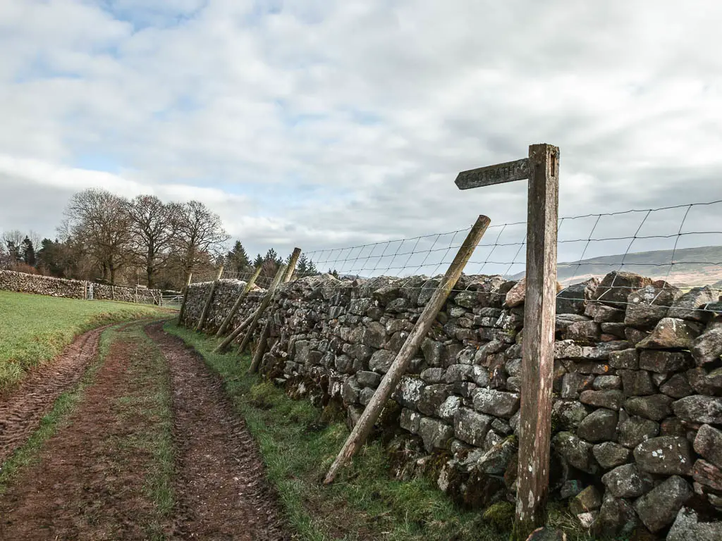 A stone wall on the right, with a dirt track running alongside it. There is a wooden trail signpost next to the wall, pointing ahead.