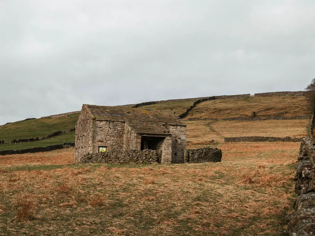 A stone shed in a field of dead grass, with a hill behind.