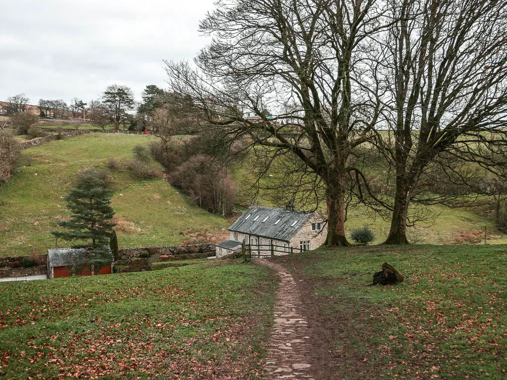 A narrow stone trail leading g downhill through the grass, towards a stone cottage at the bottom of the hill. There are a couple of big leafless trees in the field.