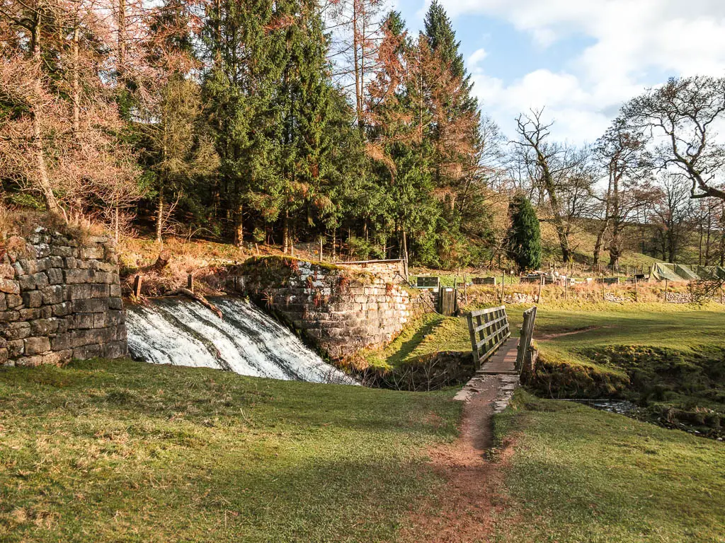 A small water fall on the left, with a wooden bridge running across the river on the right, leading from one field to another.