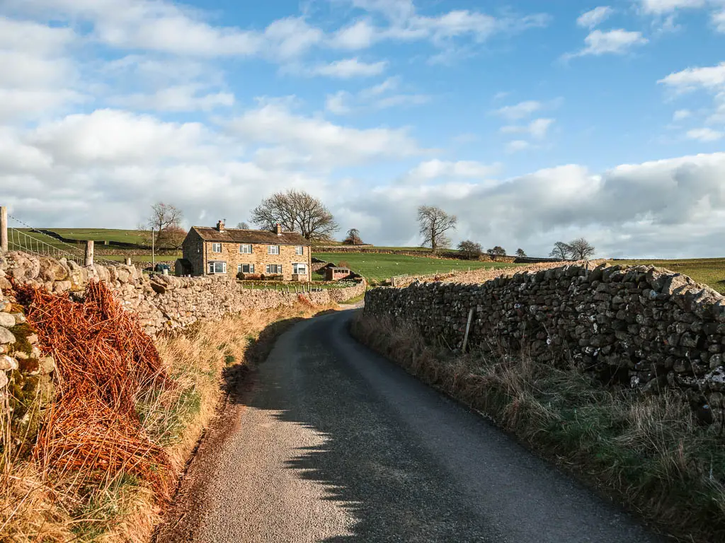 Ac outcry road lined with stone walls, and a stone walled house ahead on the left, on the walk back to Burnsall from Grassington. The sky is blue with white clouds.