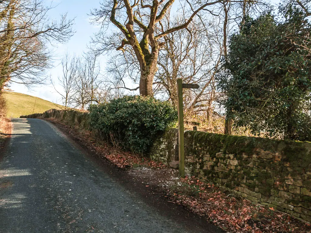 A road on the left, and a wall on the right, with a wooden trail signpost pointing to ab gap in the wall.