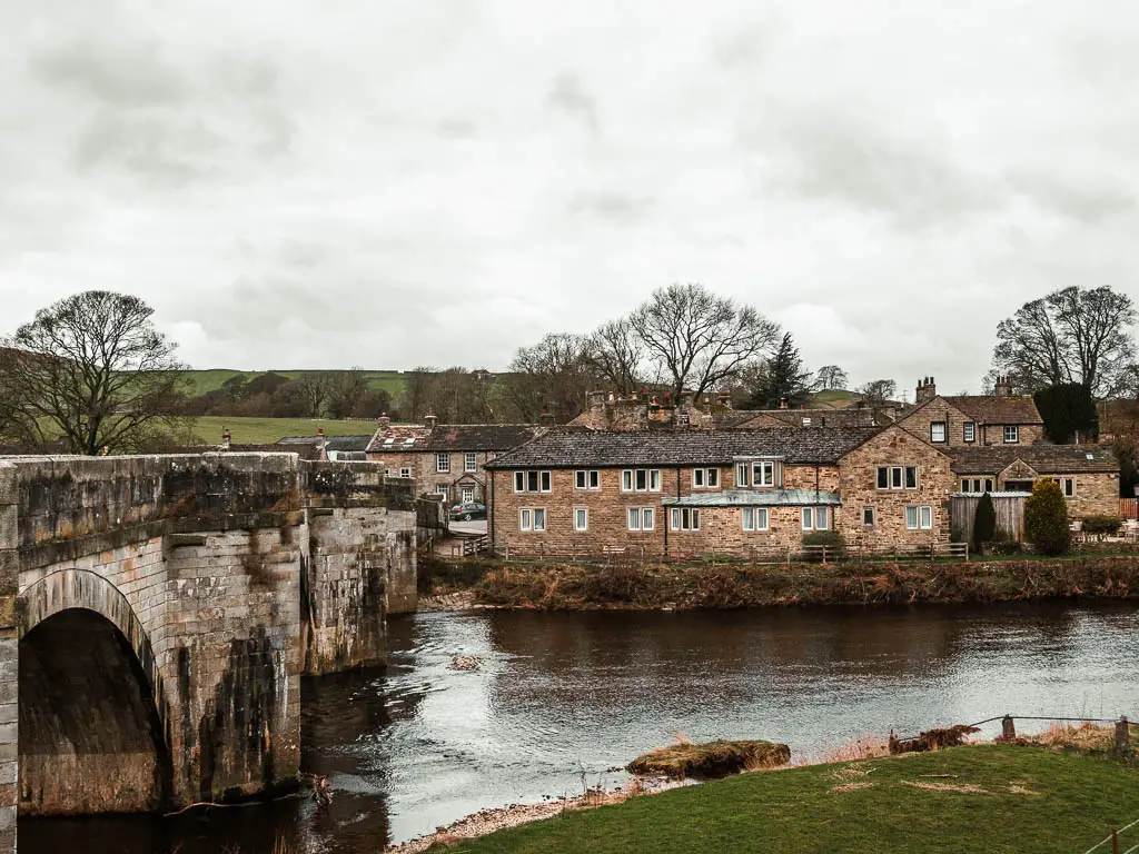 An archway bridge on the left running across the river Wye, with stone buildings of Burnsall on the other side.