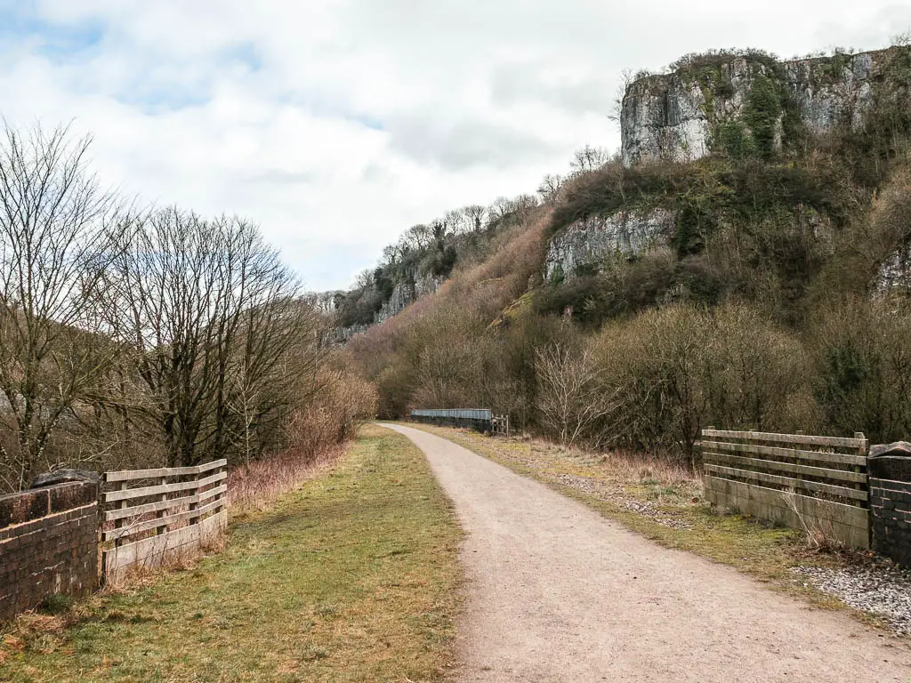 A wide path with wooden fences on weather side, and the limestone cliffs covered in some trees ahead to the right.