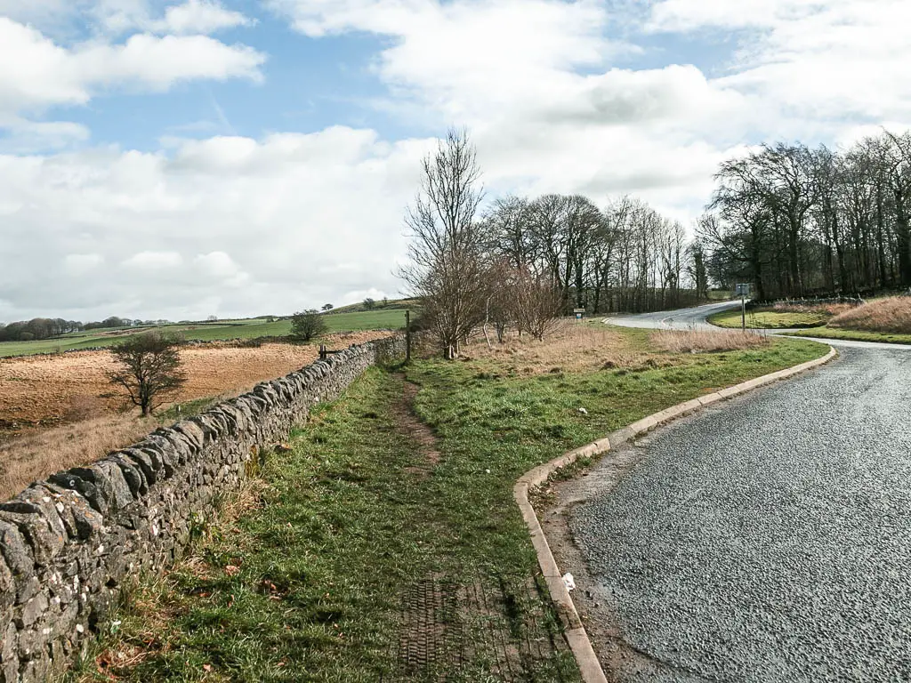 The road layby on the right, with a grass bank on the left, and a stone wall to the left of it.