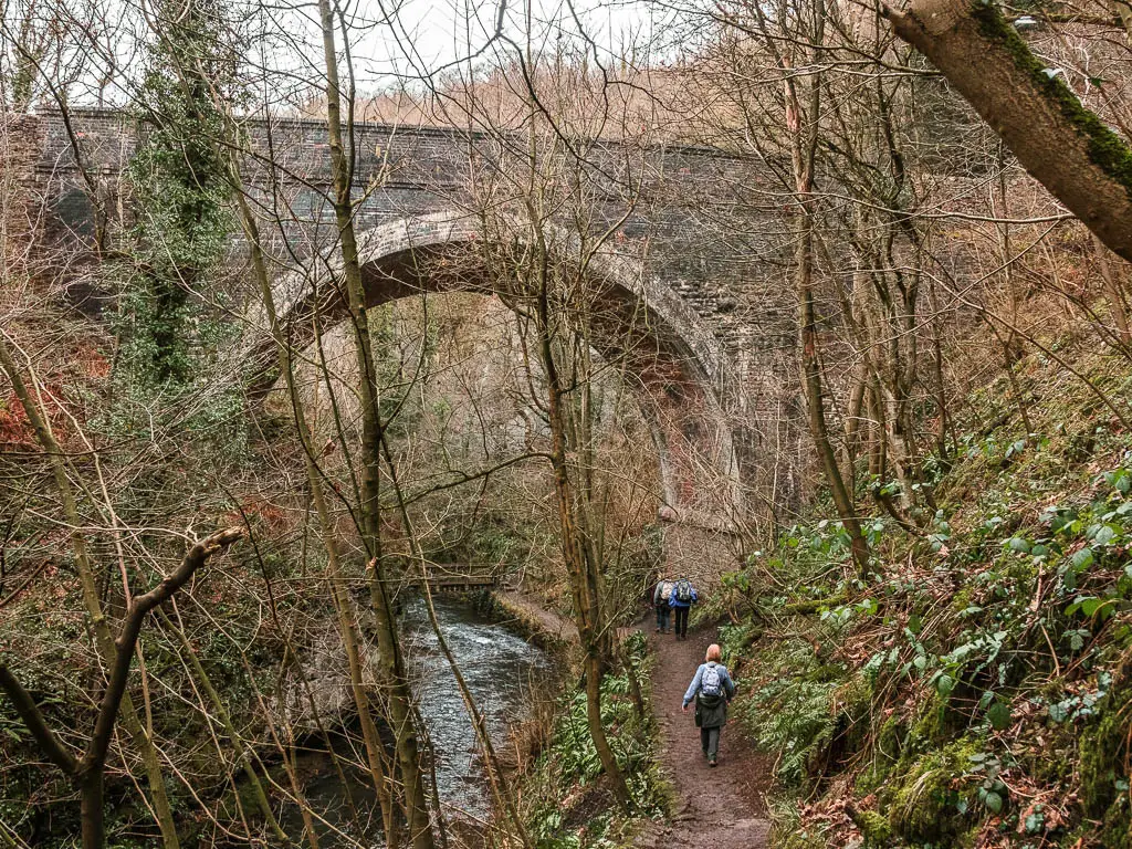 Looking down to a dirt trial with people walking along it through Chee Dale. The path leads under the archway of a bridge across the river. There are lots of straggly leafless trees.