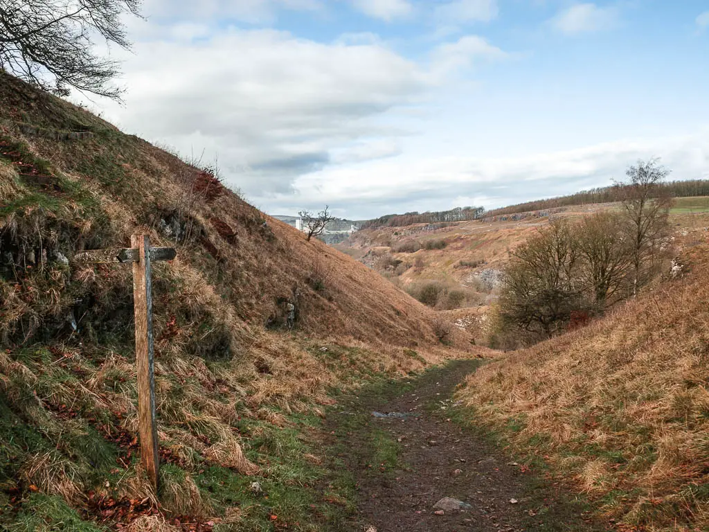 A dirt trail running through the middle of two hill slopes, with a wooden trail signpost on the left. The sky is blue with a few white clouds.