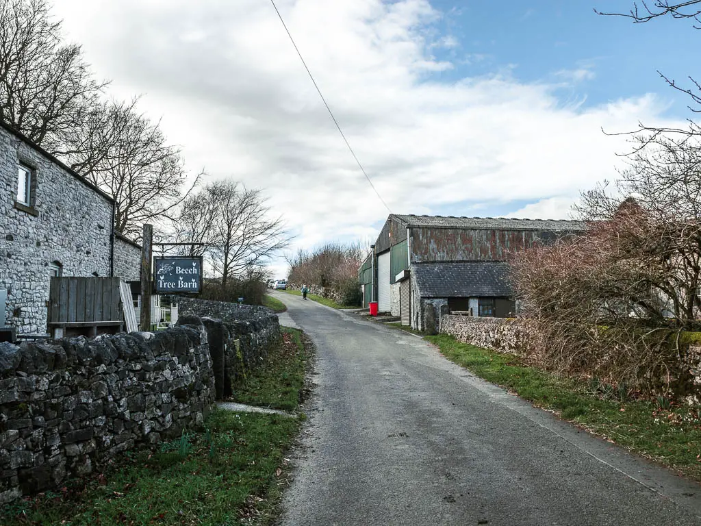 A road leading uphill, with a stone wall and stone building on the left, and large farm building ahead on the right.