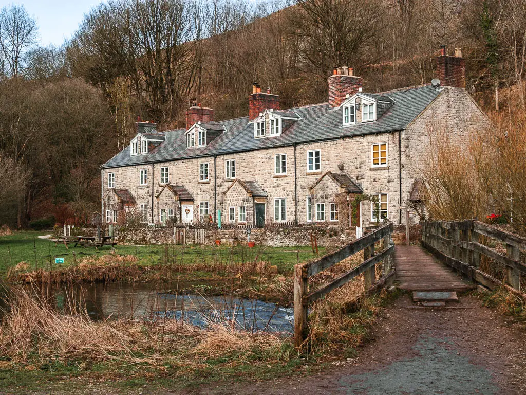 A stone walled cottage on the other side of the river, with a wooden bridge leading to it, at the start of the Chee Dale walking trail.