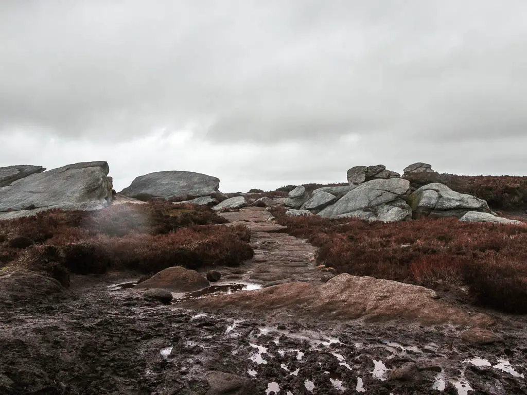 A muddy path of ground with a stone paved path on the other side, leading through some rocks when walking along Derwent Edge.