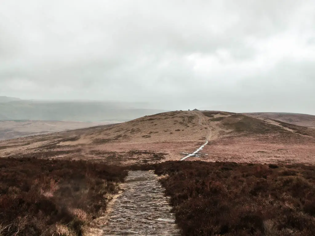 A stone and dirt trail leading ahead to a hill across the moors on a misty day.