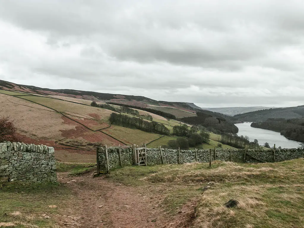 A dirt trail leading through a stone wall on the left, on the walk down into the valley, with Derwent Edge to the left and the reservoir below. 