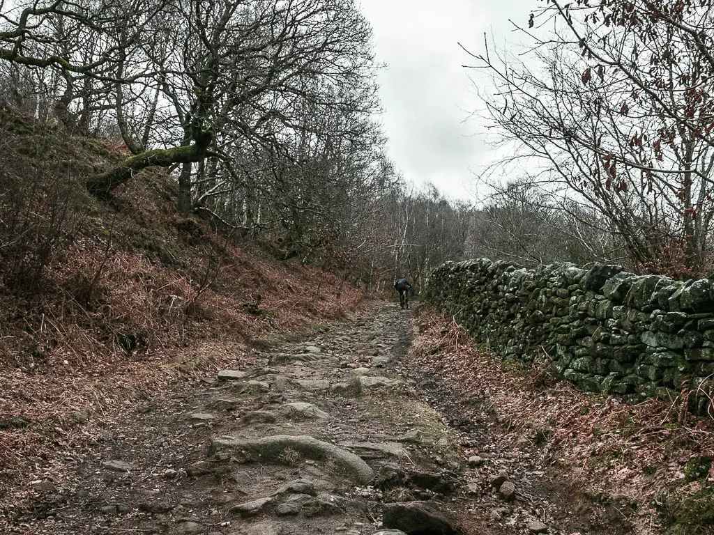 A rocky trail leading uphill, with a stone wall on the right. there is a person going up the hill on a bike.