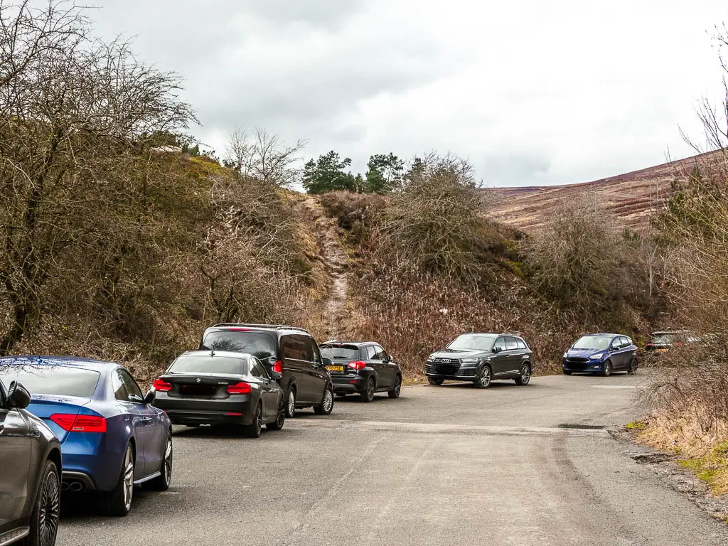 A lay-by car park, with cars parked along the side, and a hill and trail ahead.