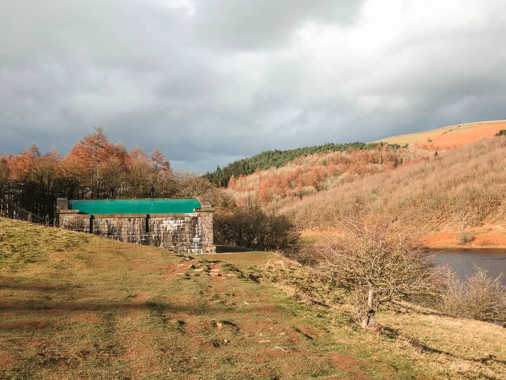 A brick shed building with a green roof, with trees poking up from the other side. There are grey and white clouds in the sky.