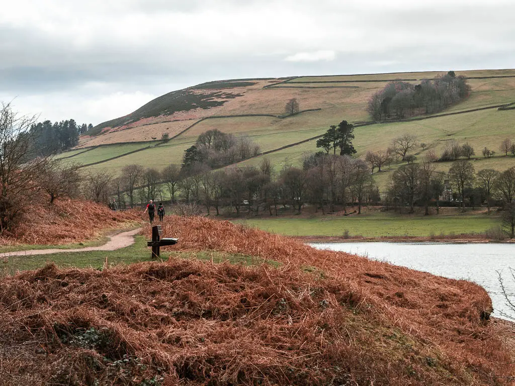 A wooden bench facing the Ladybower Reservoir, and a couple of people walking along the trail in the distance. There is a big hill with green grasses fields and a few trees on the other side of the water.