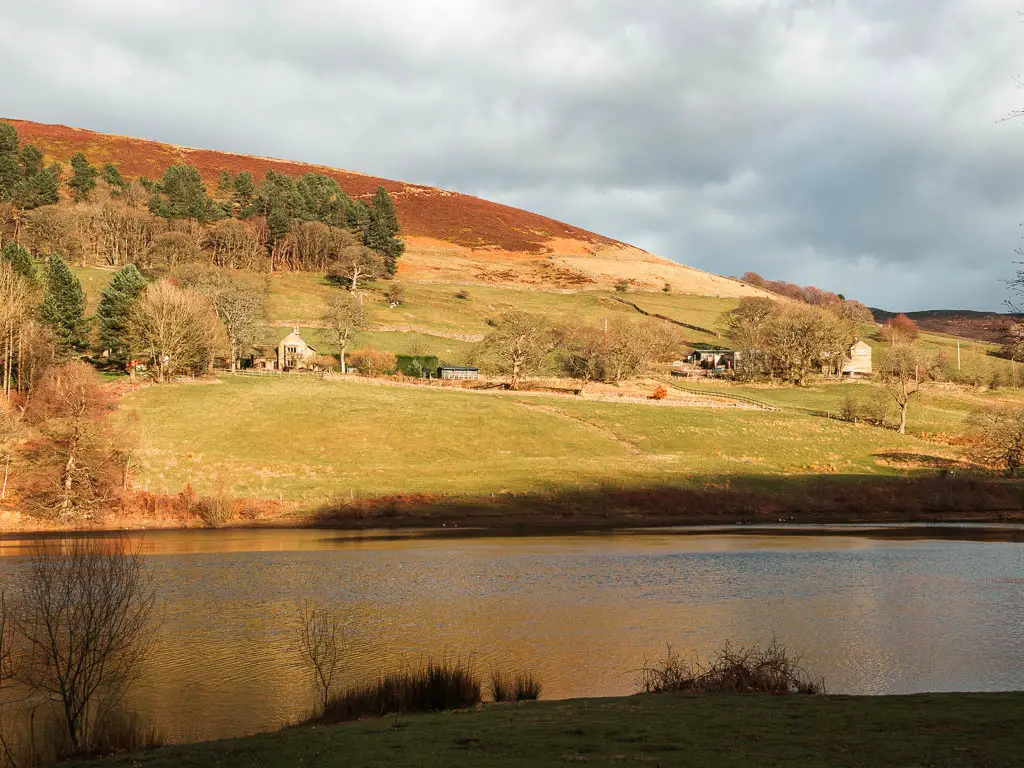 Looking across the Ladybower Reservoir, to a hill on the other side on the north end of the walk. There are a few houses on the hill, and the sun is shining down brightly on it.