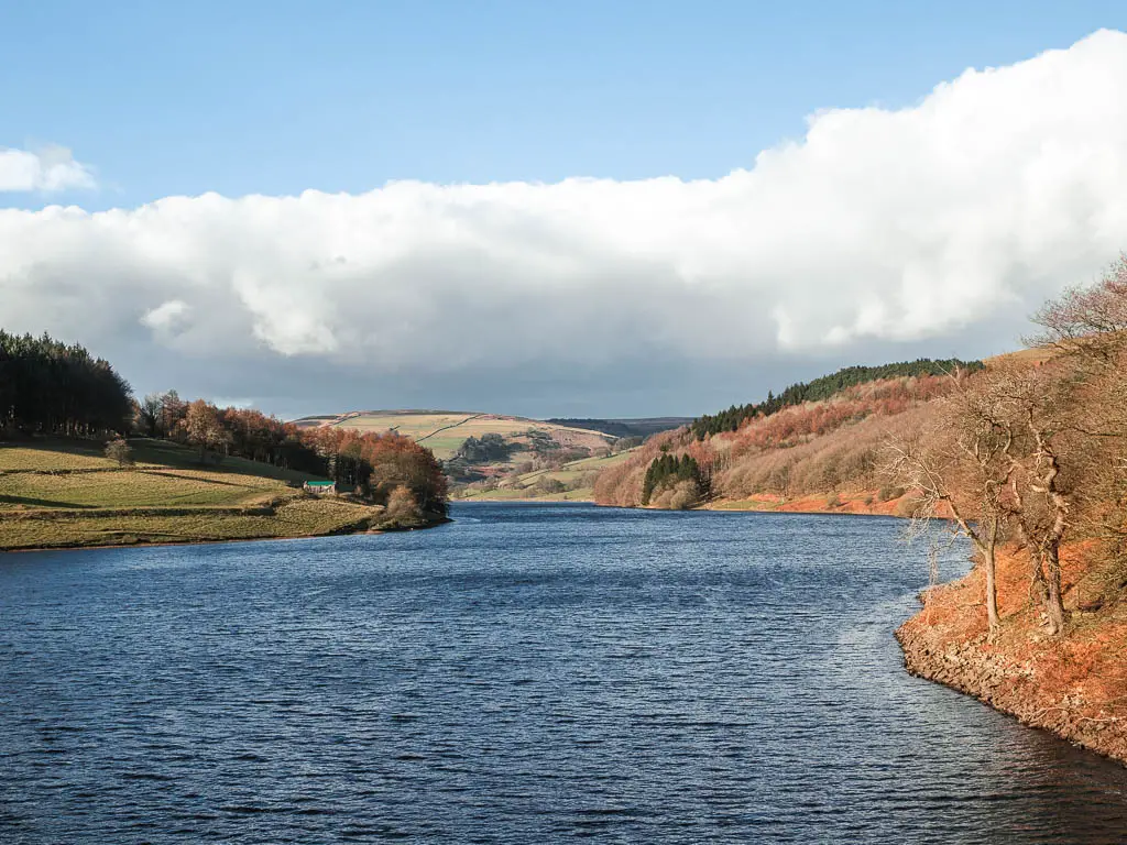 Looking across the rich blue water of the Ladybower Reservoir, at the start of the walk around it. The reservoir is surround by undulating hills with trees, in shades of orange, green, and brown.