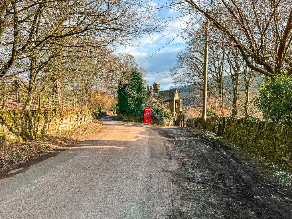 A country road, lines with stone walls, and a house ahead with a red telephone box outside.