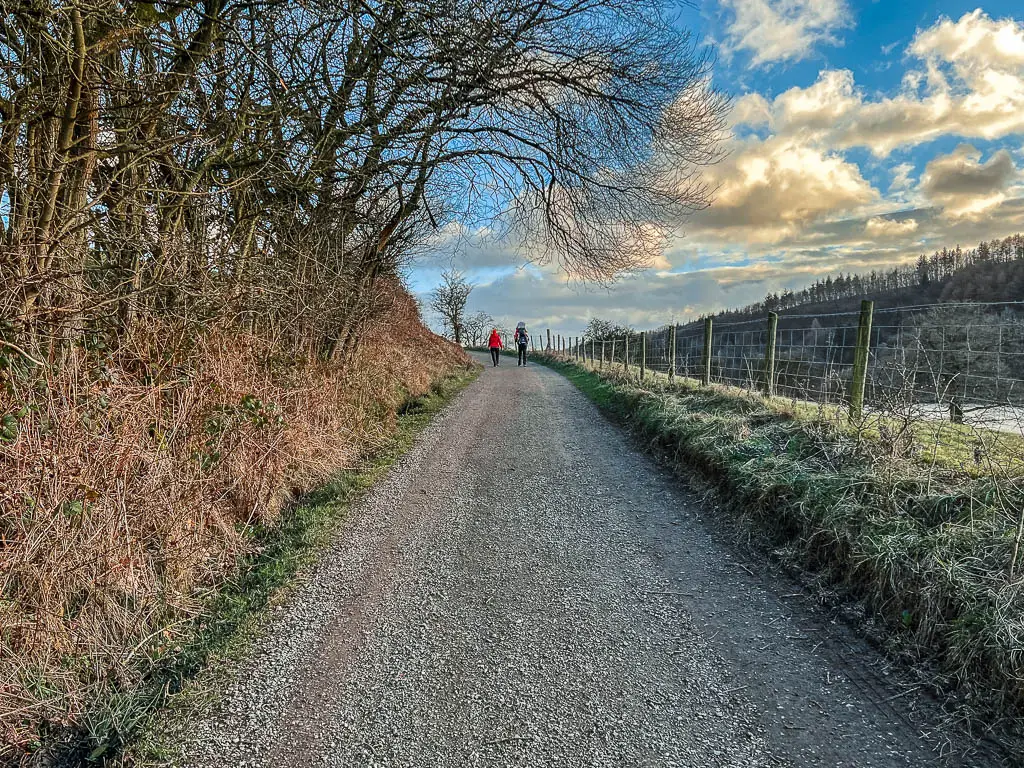 A gravel path leading uphill, with a couple of people walking up it. There is a metal fence on the right and scraggly bushes and trees on the left.