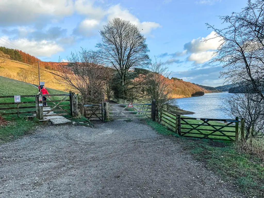 The gravel trail leading to three wooden gates, with two people walking through the left gate, and a view to the Ladybower Reservoir on the right.