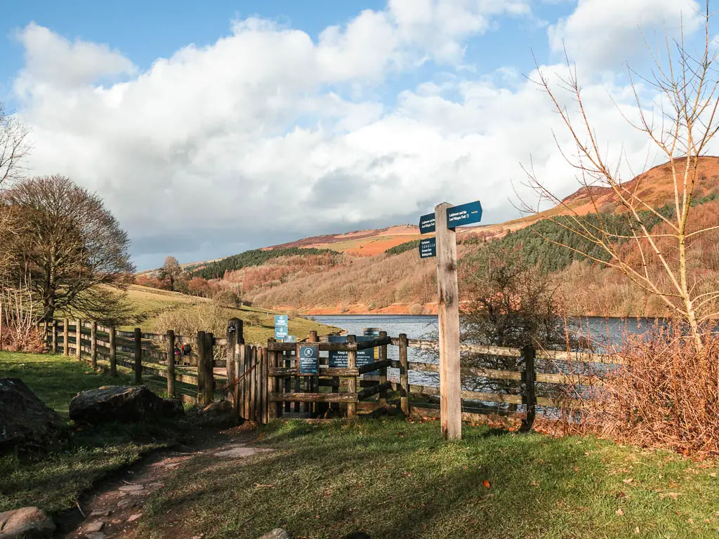 A wooden fence and gate, with a wooden trail signpost marking the start of the walk around the Ladybower Reservoir. There is an undulating hill on the other side, with a view to Derwent Edge.