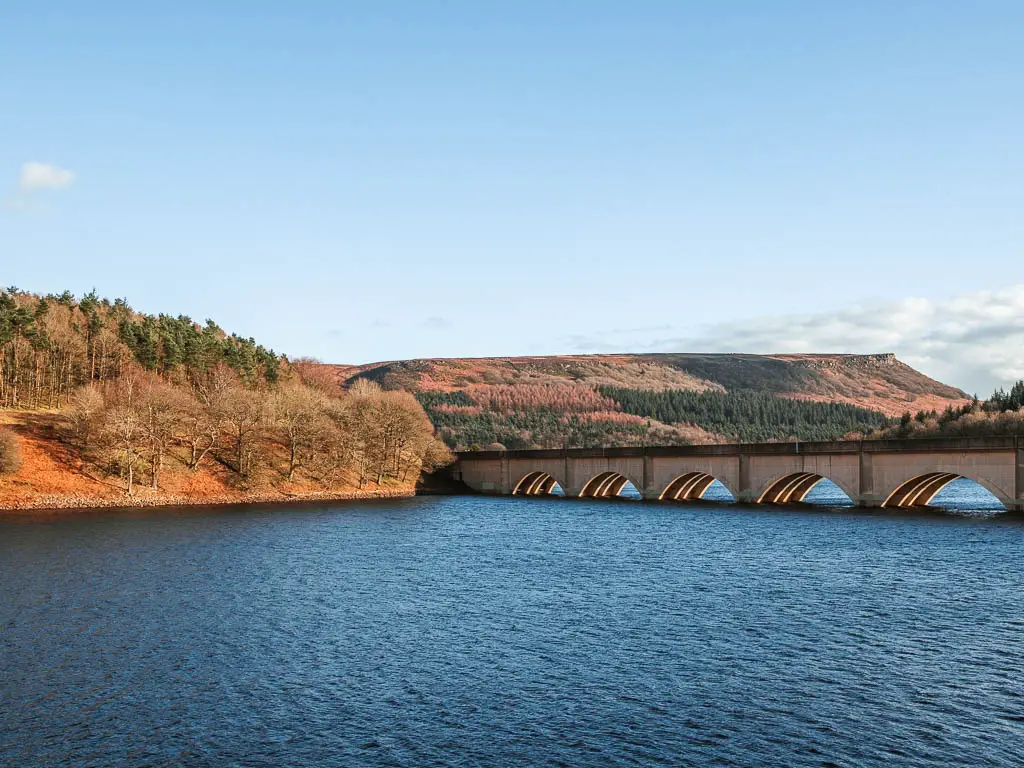 The Ashropton viaduct running over the blue water of the Ladybower Reservoir, with woodland tree covered hills on the other side.