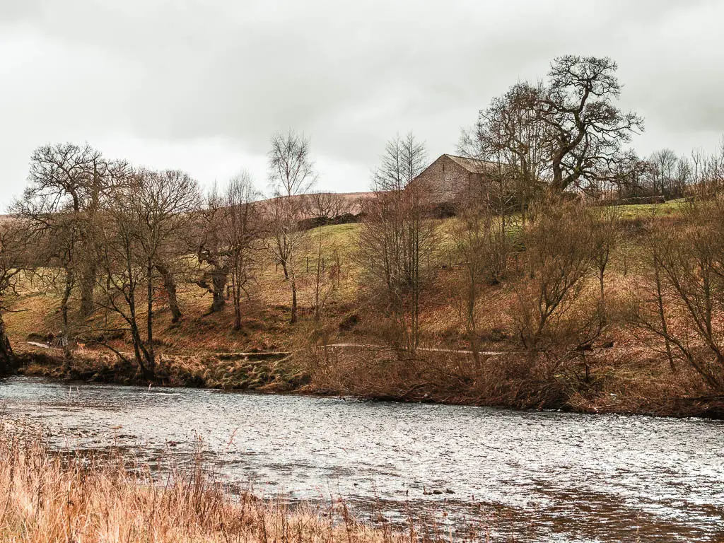 Looking across the river, to a hill on the other side, with a stone shed on top, partially obscured by leafless trees.