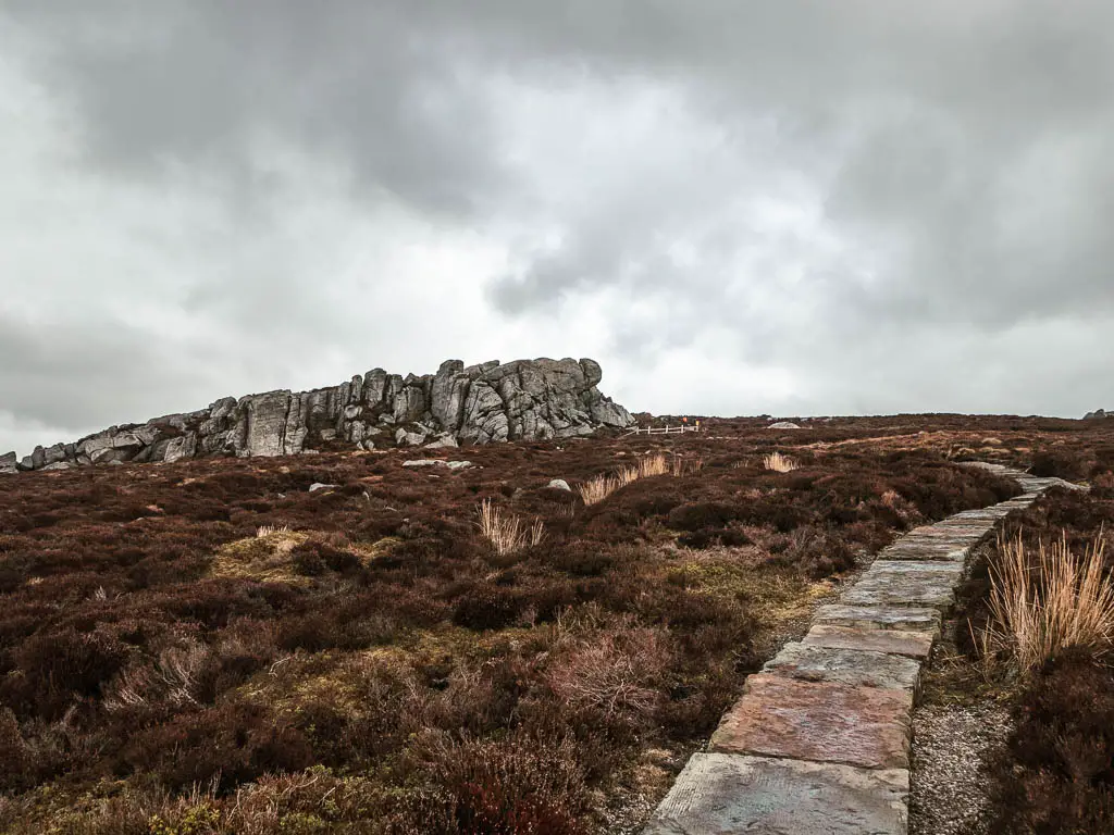 A stone paved trail on the right, and the big rocky outcrop of Simon's Seat ahead to the left, on the walk to the top. The ground is covered in out of bloom heather.