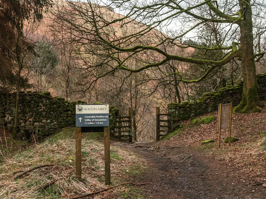 A signpost pointing the way to Botlon Abbey and the Valley of Desolation, on the walk down from Simon's Seat. The sign points through a wooden gate in the stone wall, with straggly woodland on the other side.
