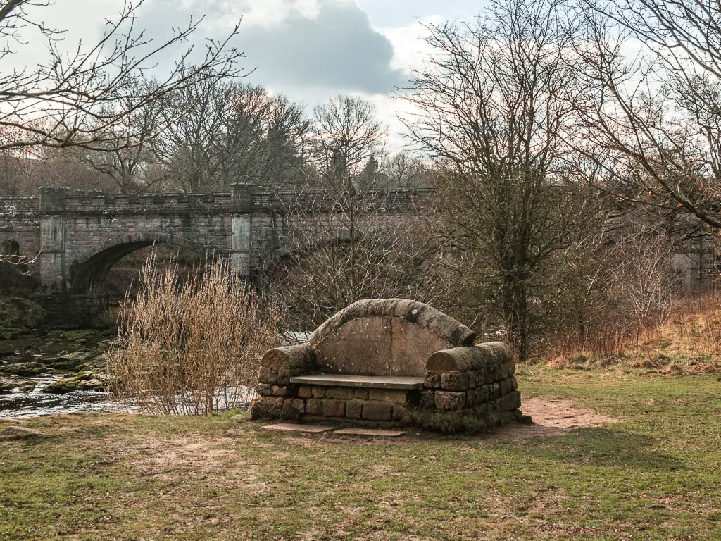 A stone seat on the gras bank next to the river. surround by leafless trees, and a bridge in the background.