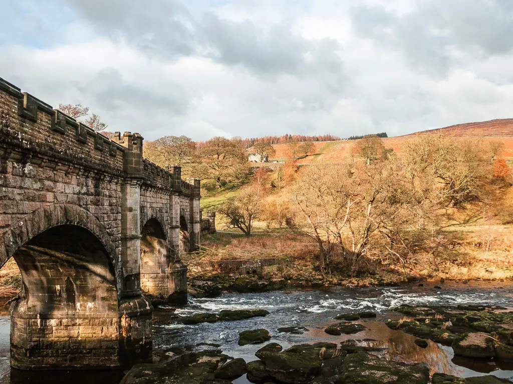 A large stone bridge on the left, running across the river, with the sun shining down on the hills on the other side, at the end of the Simon's Seat walk.