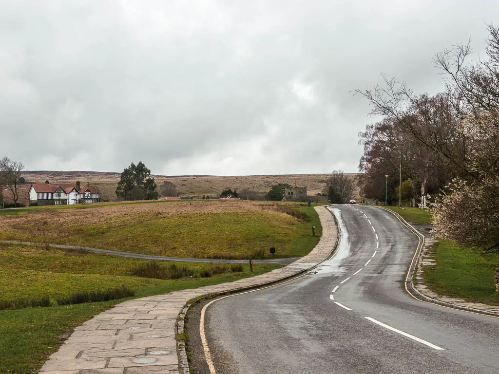 The road snaking ahead, with a grass bank on the left side, and a cottage oil the distance to the left. 