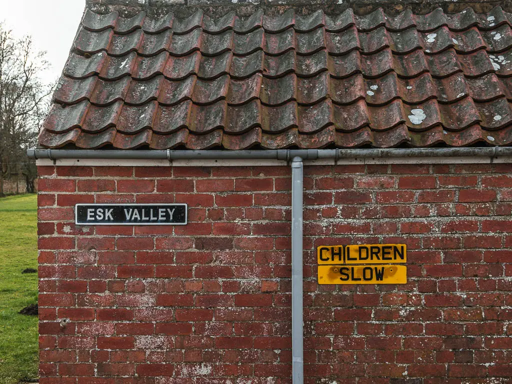 A brick wall, with a black sign saying 'Eske valley', and yellow sign saying 'children slow', along the rail trail walk from Goathland to Grosmont