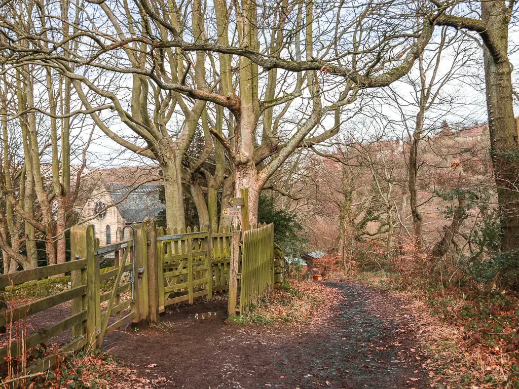 A dirt path leading downhill to the right, and a wooden fence and gate on the left side, with a view to part of the church past the gate.