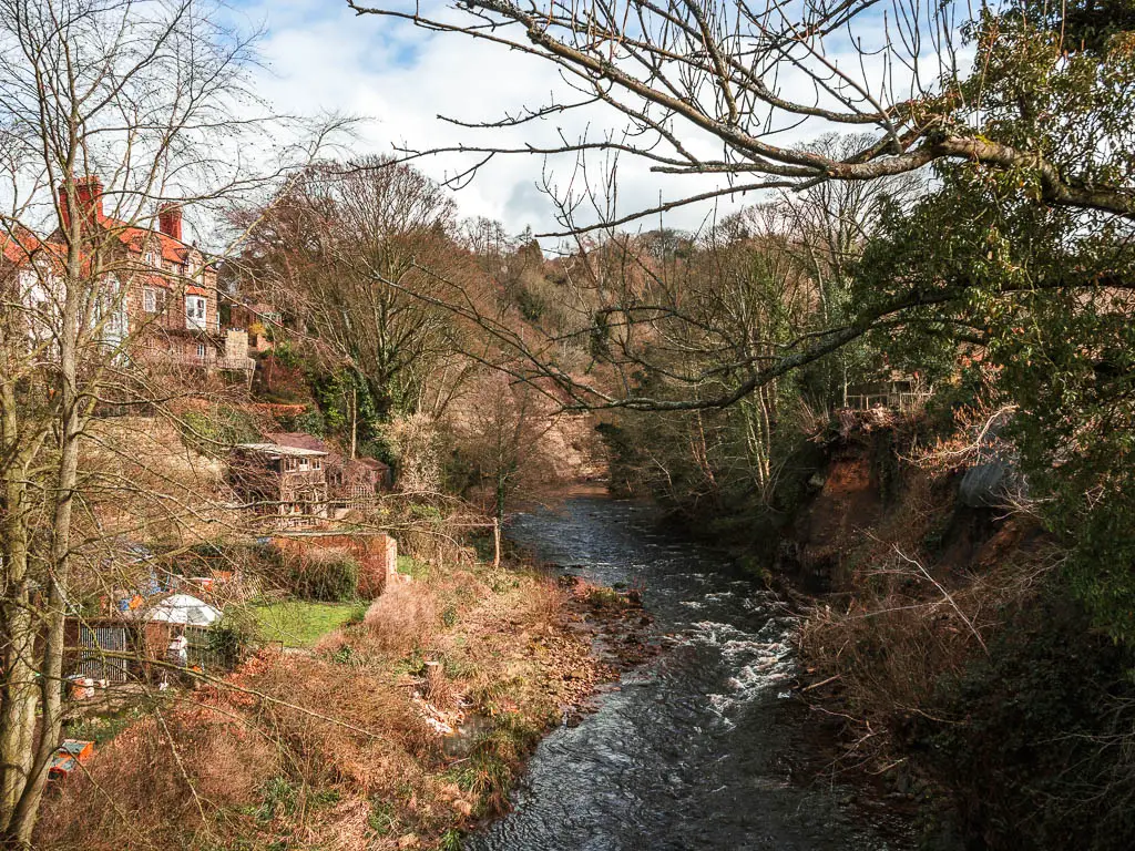 The river flowing between the banks, and a house up the hill on the left, when walking through Grosmont. There are lots of bushes and trees along the river bank side.