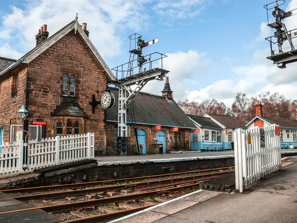 Looking across the railway tracks of Grosmont station, to the platform on the other side, and the hut buildings in blue and white. there is a big clock face on the side of the brick building. There are white picket fences. 