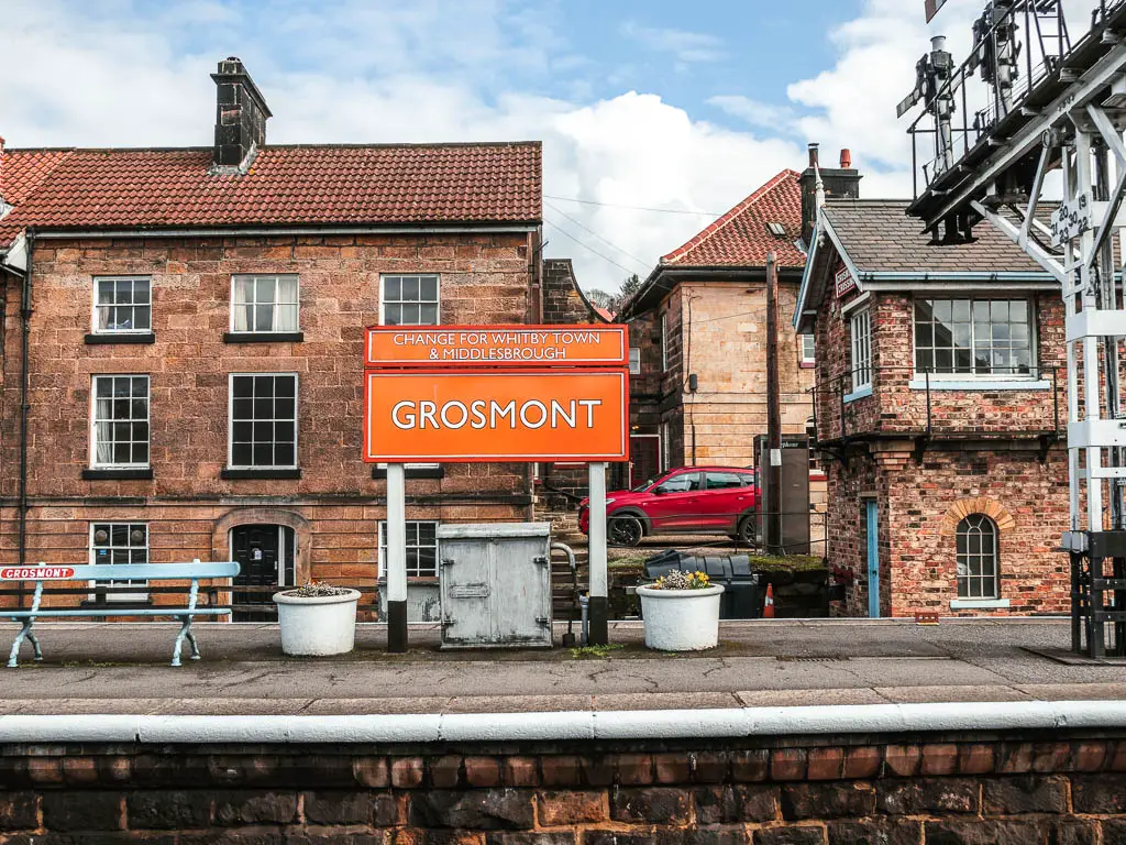 An orange sign saying 'Grosmont' on the platform of the railway station. There is a light blue bench on the left of the sign. There are street houses behind.