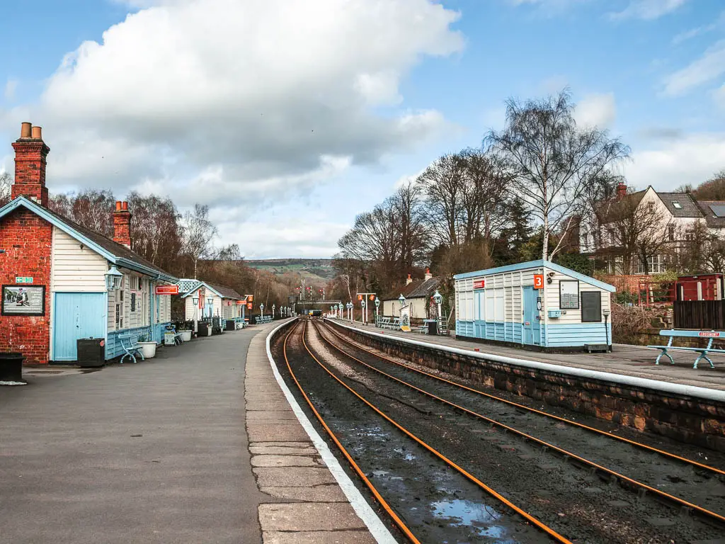 Standing on the platform of Grosmont station, looking along the railway track. The platforms have small blue and white hut buildings, with orange hanging signs.