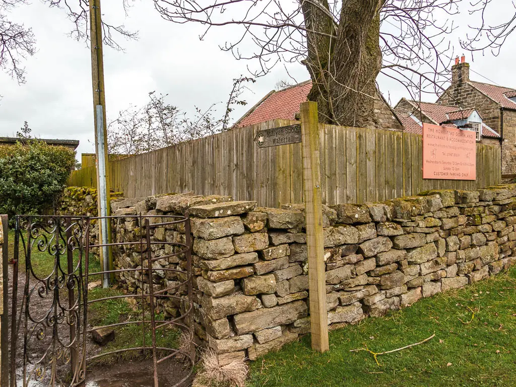 A wooden trail sign next to a stone wall, pointing the way to walk through a metal gate, to get to Mallyan Spout waterfall.