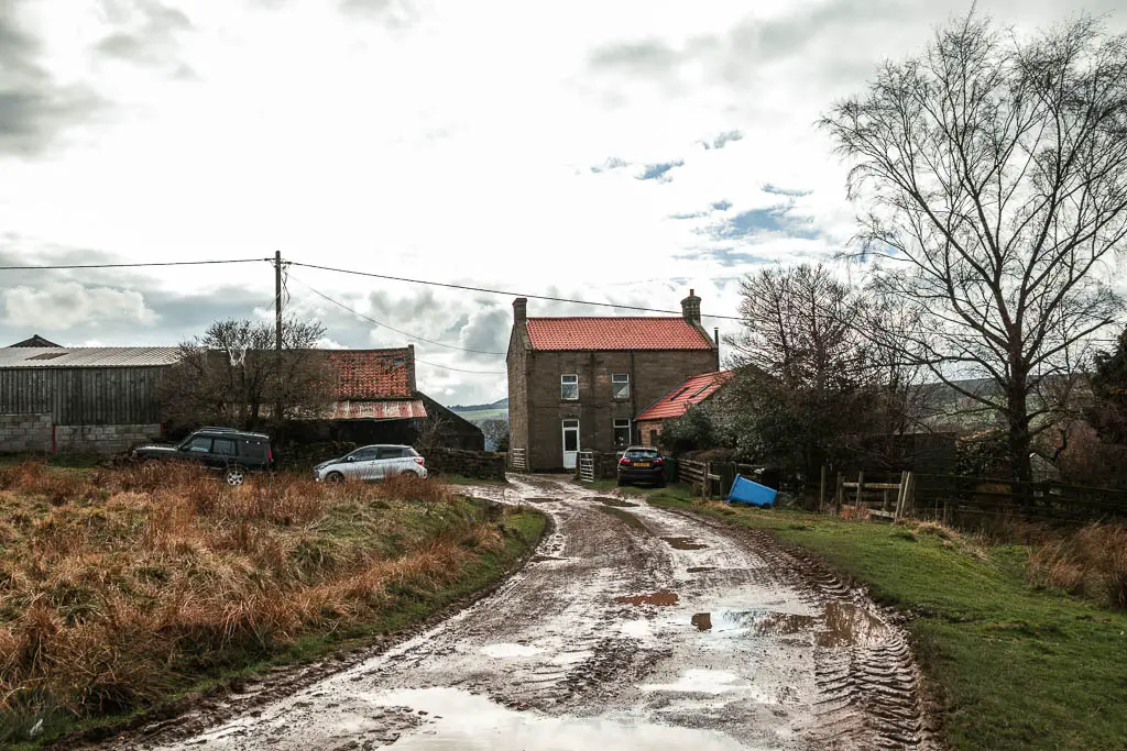 a muddy, wet road leading straight towards some farm houses, with some cars parked outside.