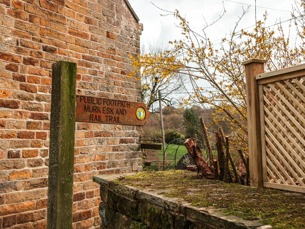 A wooden trail signpost, infant of a brick wall, pointing right to walk along the rail trail. 