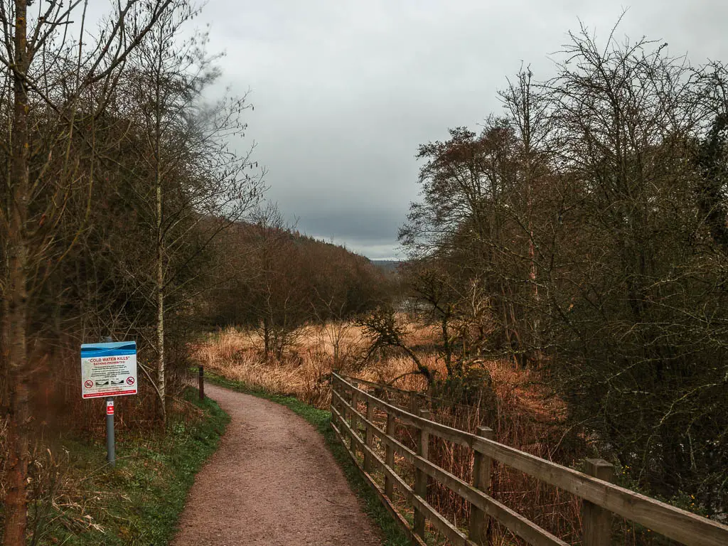 A path with a wooden railing on the right. It is lined with leafless bushes and trees. There is a sign on the left side of the path.