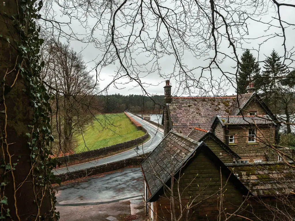 Looking down over the stone walled cottage, towards the road bridge. There is a grass hill field on the left side of the road. There are leafless tree branches hanging from above.