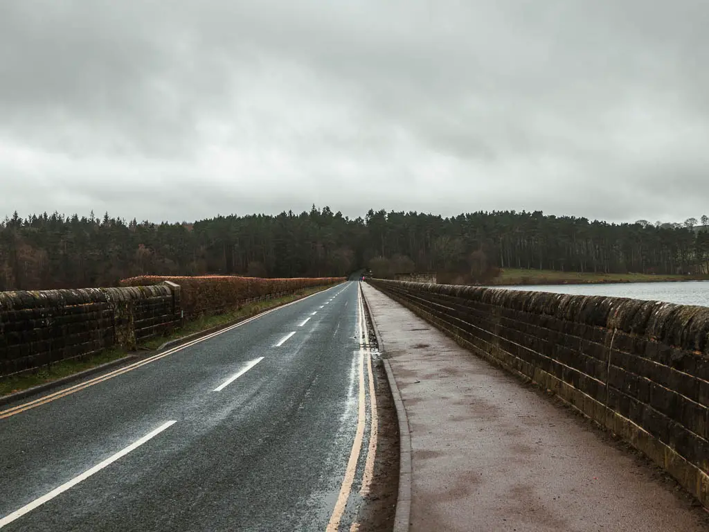 Looking along the road bridge with a pavement on the right. There is a stone wall on the right side of the road and hedge on the left side. There is a mass of woodland trees on the other end of the road.