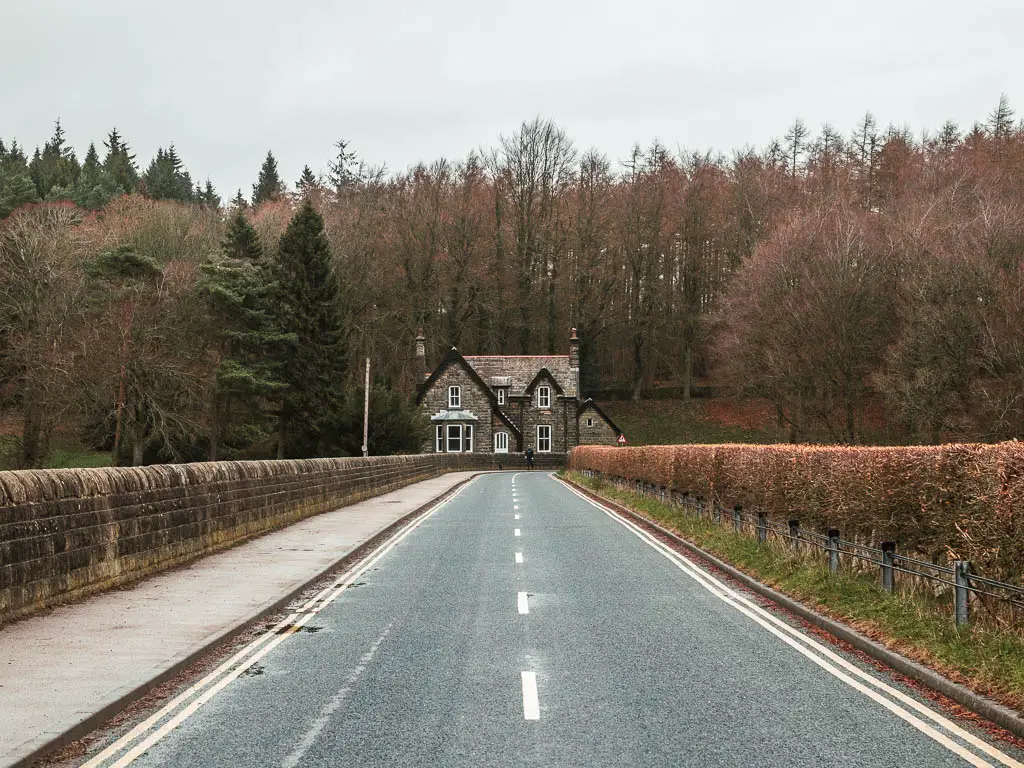 A road bridge leading to a stone cottage, surround by woodland trees. There is a pavement and stone wall on the left side of the road, and hedge on the right side.