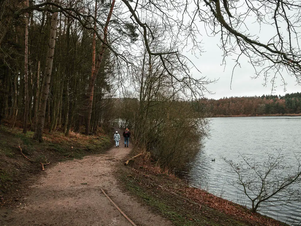 The dirt trail curving along the left side of the Fewston Reservoir, with a couple of people walking along it. The left side of the trail is full of woodland trees.