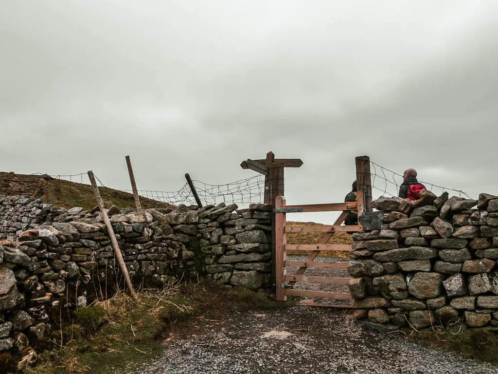 A wooden gate in a stone wall, with a wooden trail signpost on the other side. There are a people standing on the other side of the wall.