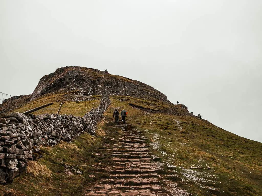 Stone steps leading uphill along the grass covered hillside, with the summit of Pen-y-Ghent ahead, on the walk up to the top. There are a couple of people walking up the steps ahead. There is a stone wall on the left.