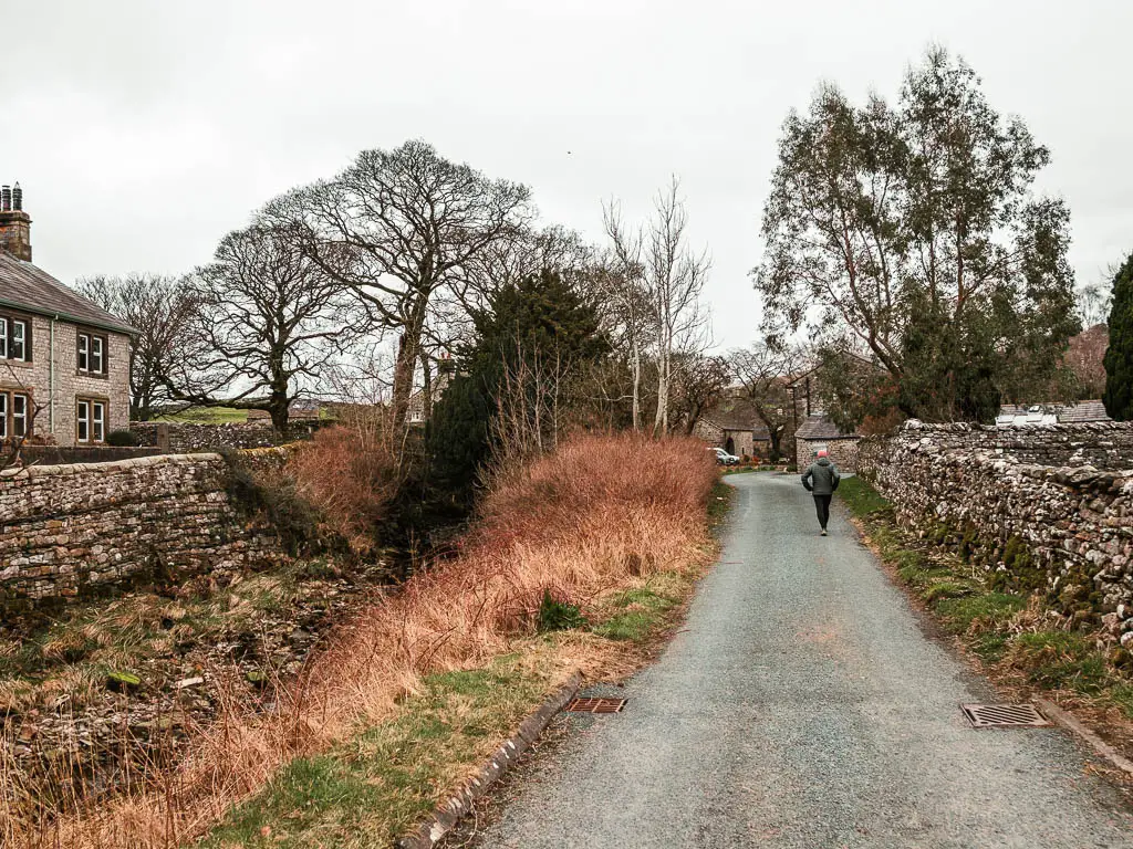 A road with a stone wall on the right, grass and hay on the left, and a house to the left of that behind a stone wall. There is a man walking on the road.